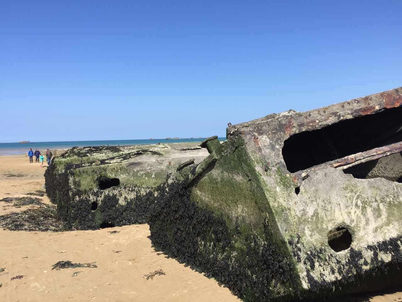 Rovine del porto artificiale Mulberry Harbour sulla spiaggia di Arromanches, Normandia, con persone che camminano sulla sabbia in lontananza