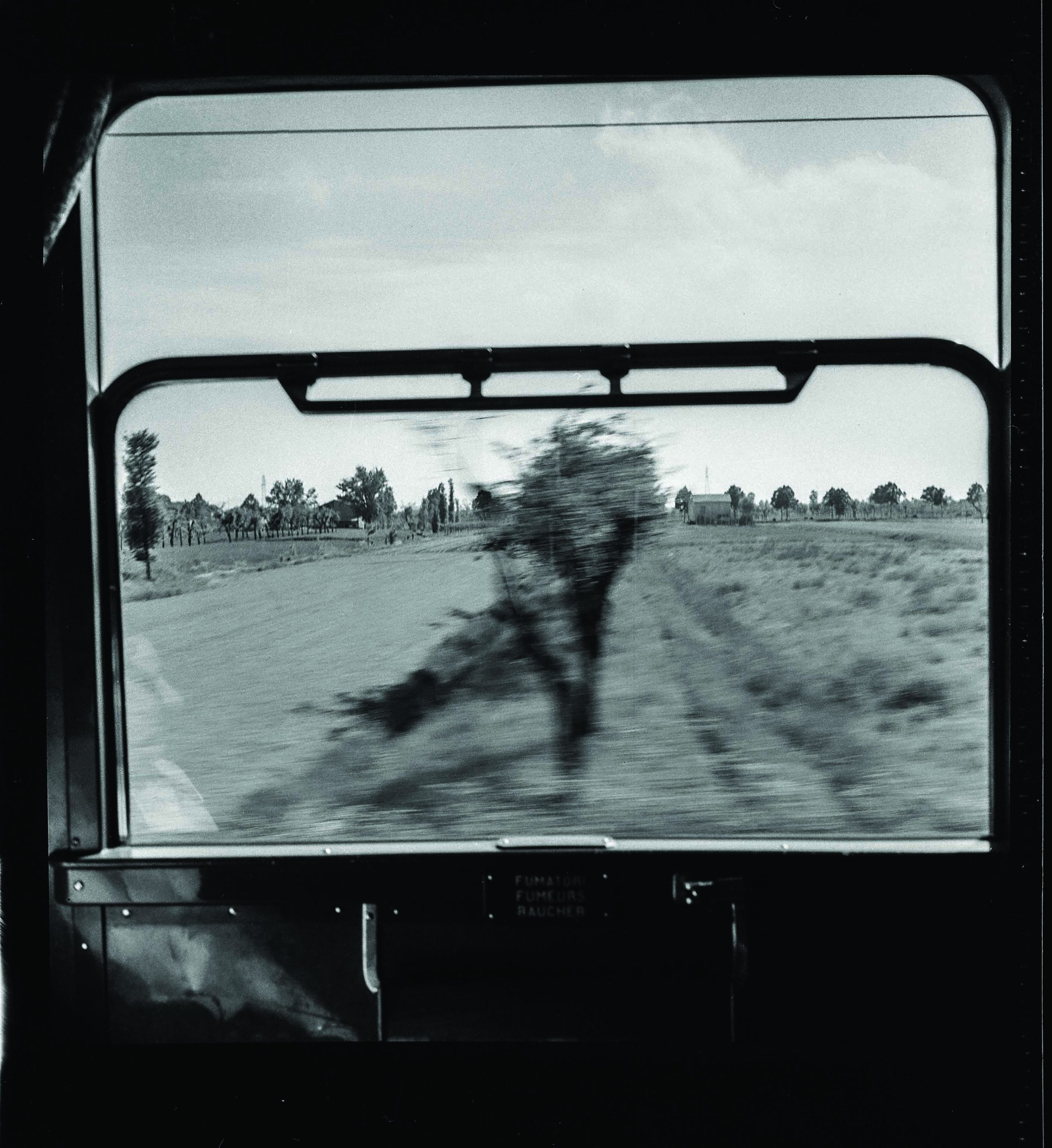 Fotografia in bianco e nero scattata dall'interno di un treno in movimento, mostrando il paesaggio rurale attraverso una finestra aperta. L'immagine cattura l'effetto sfocato del movimento, con alberi e campi che scorrono rapidamente.