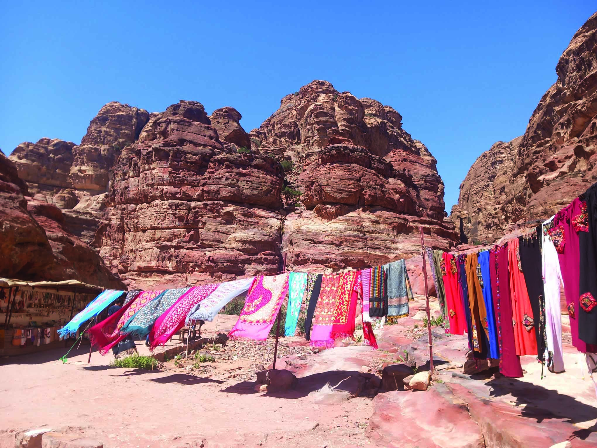 Scena in un mercato all'aperto con sciarpe colorate appese ad asciugare, situato tra formazioni rocciose rosse e imponenti, sotto un cielo azzurro. Ambiente desertico e vivace.