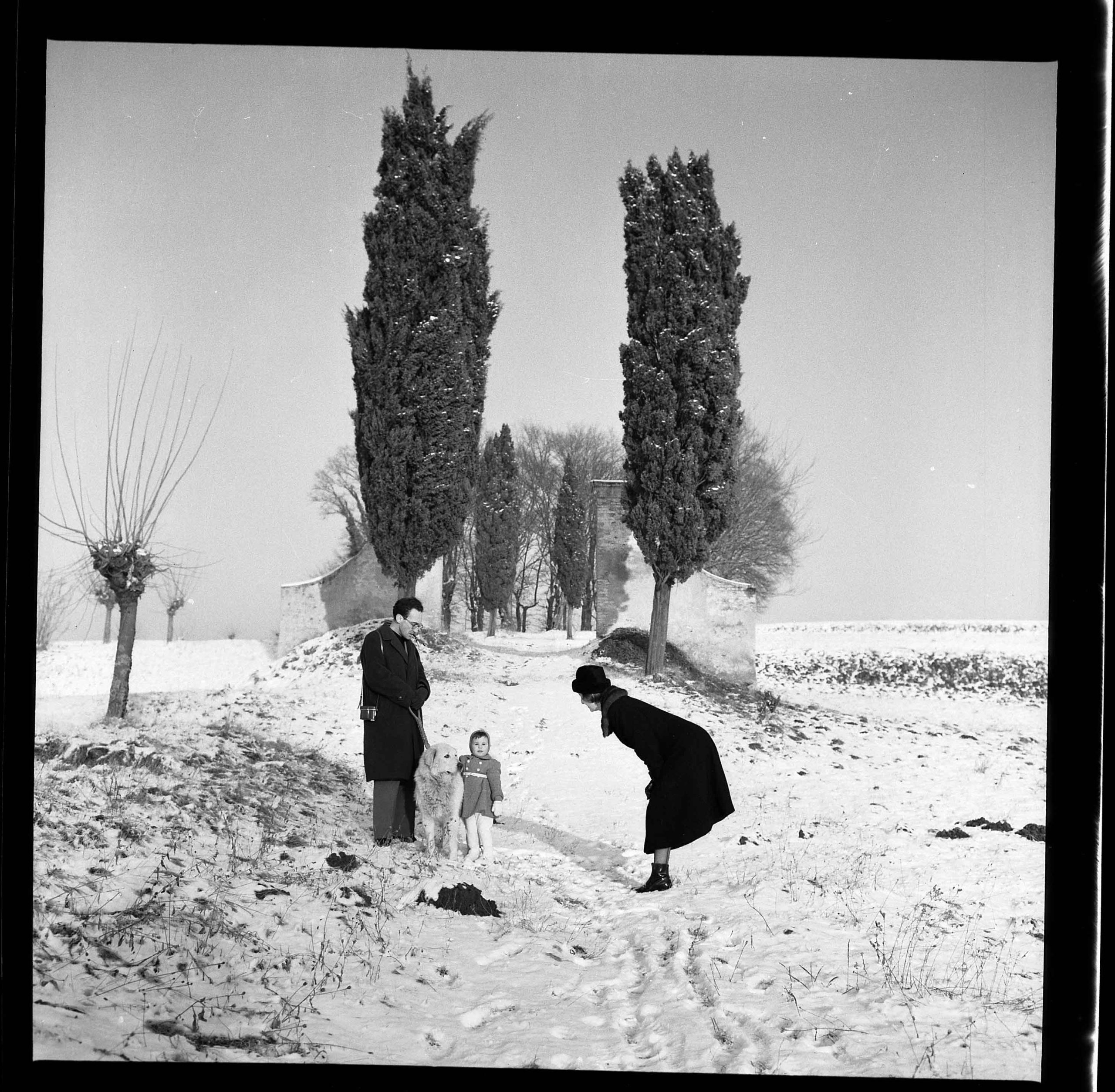 Fotografia in bianco e nero di un paesaggio invernale con neve. In primo piano, una famiglia composta da un uomo, una donna e una bambina, insieme a un cane, posano in un campo innevato. Sullo sfondo, un sentiero fiancheggiato da cipressi conduce a una costruzione. L'atmosfera è serena e l'immagine cattura un momento di interazione familiare.