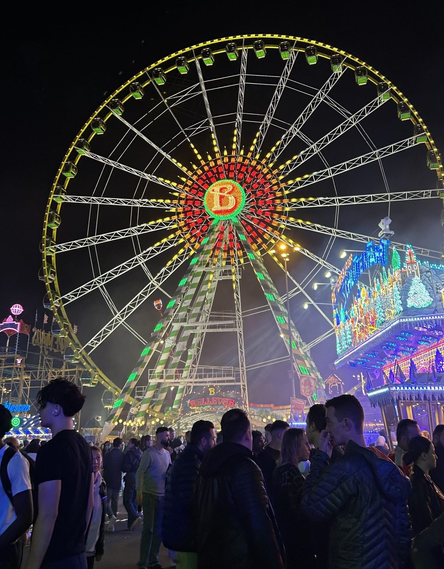 Ruota panoramica illuminata di notte, con luci colorate e una folla di persone in primo piano. Sullo sfondo si vedono altre attrazioni di una fiera, con edifici decorati e insegne luminose.