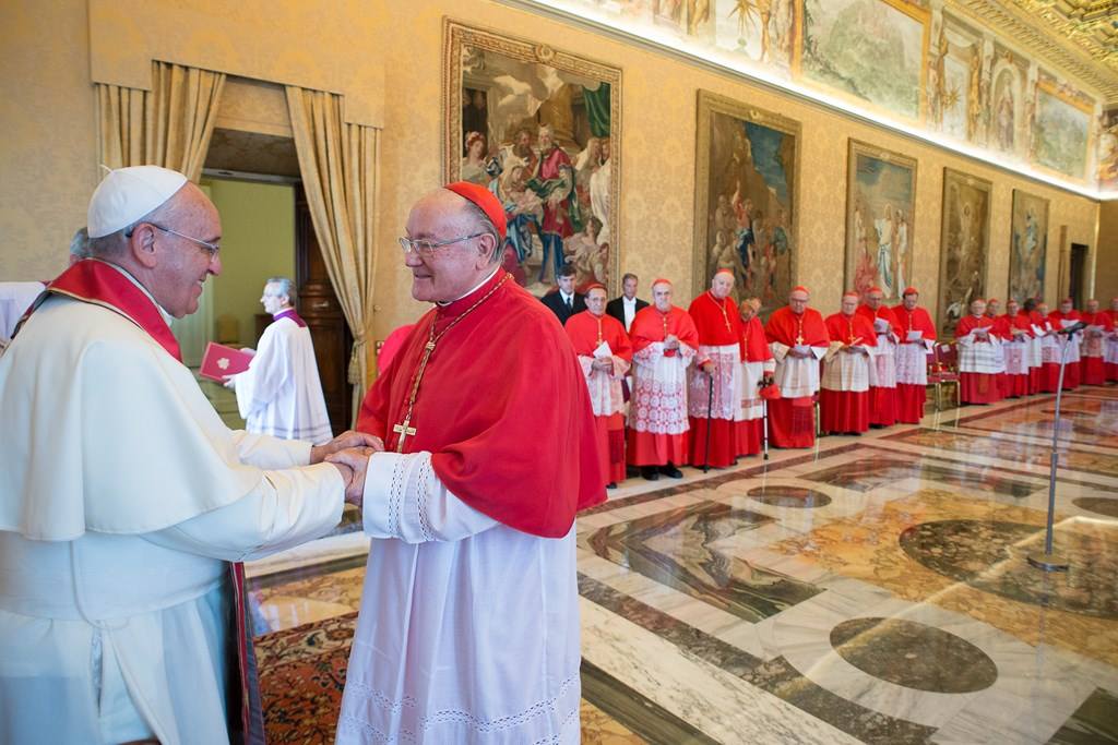 uomo vestito di bianco stringe la mani a un uomo con mantella e papalina rossa. Sullo sfondo una fila di uomini con mantella e papalina rossa